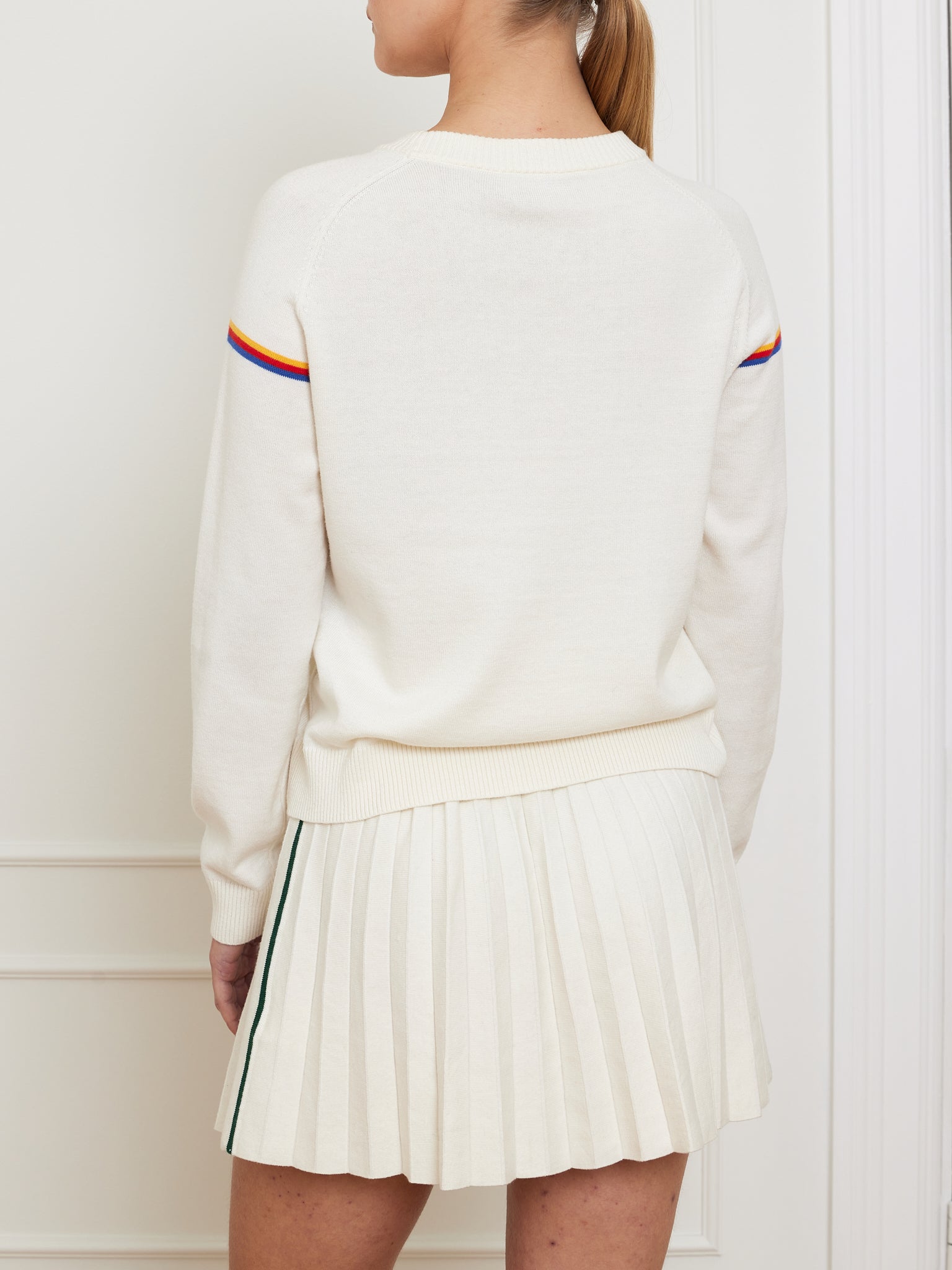 A person showcases a vintage athletic style with the We Norwegians Grand Slam Sweater Women in cream, featuring colorful sleeve stripes, perfectly paired with a pleated cream skirt. Their ponytail harmonizes with the white paneled background.