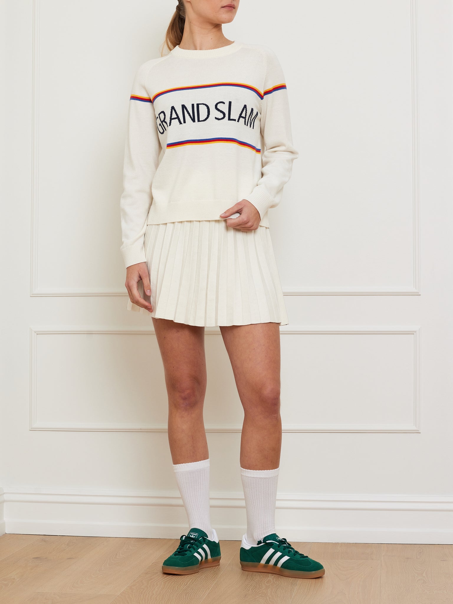 A person showcases vintage athletic style with a "Grand Slam Sweater Women" by We Norwegians in soft cream and colored stripes, paired with a pleated skirt. White knee-high socks and green sneakers complete the look against a simple white wall and floor molding backdrop.