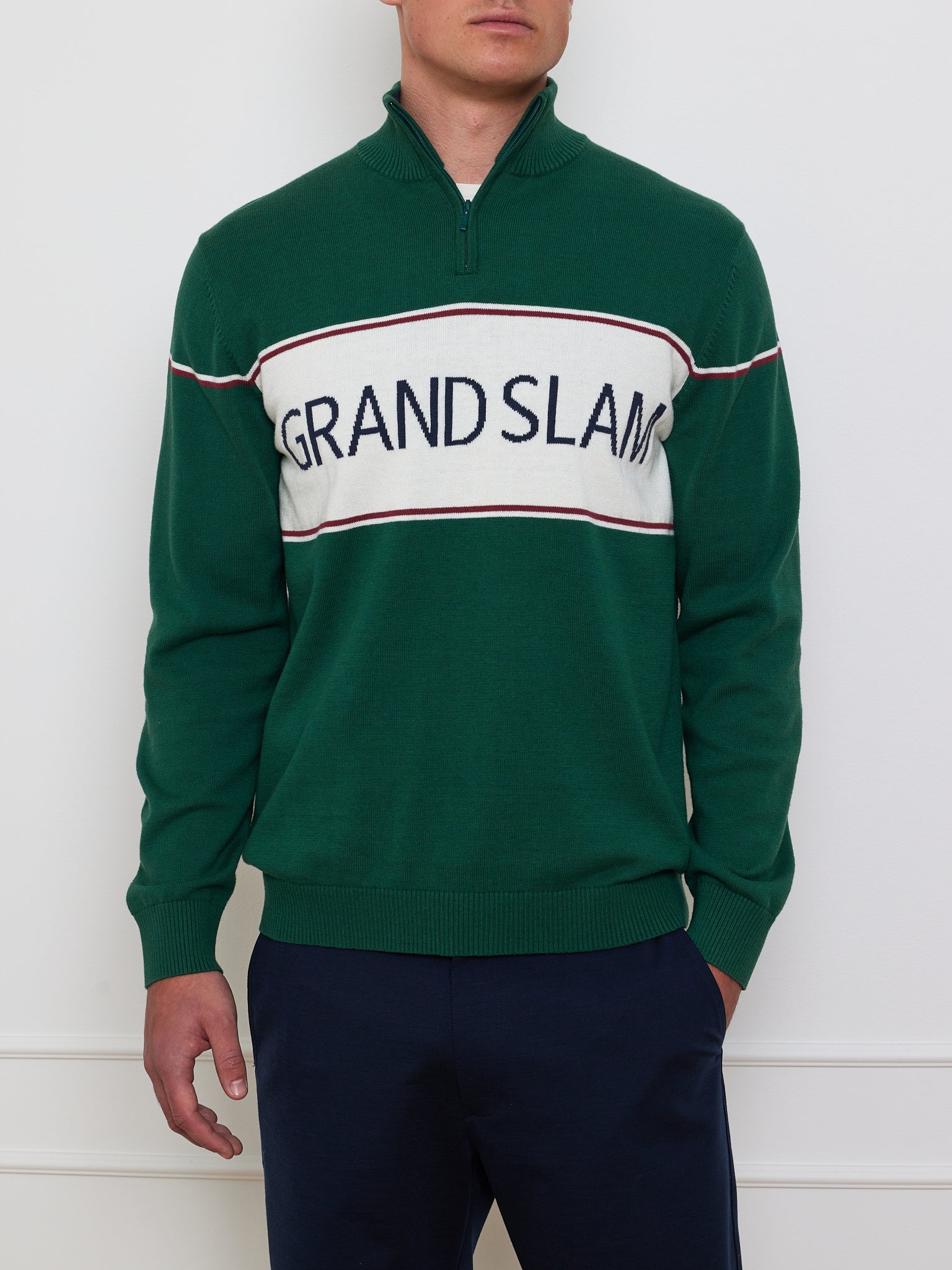 A person wears We Norwegians' Grand Slam Sweater Men, featuring a green cotton-merino blend with a quarter-zip and bold "GRAND SLAM" text over a white stripe. This vintage athletic look is paired with dark pants against a light-colored wall.