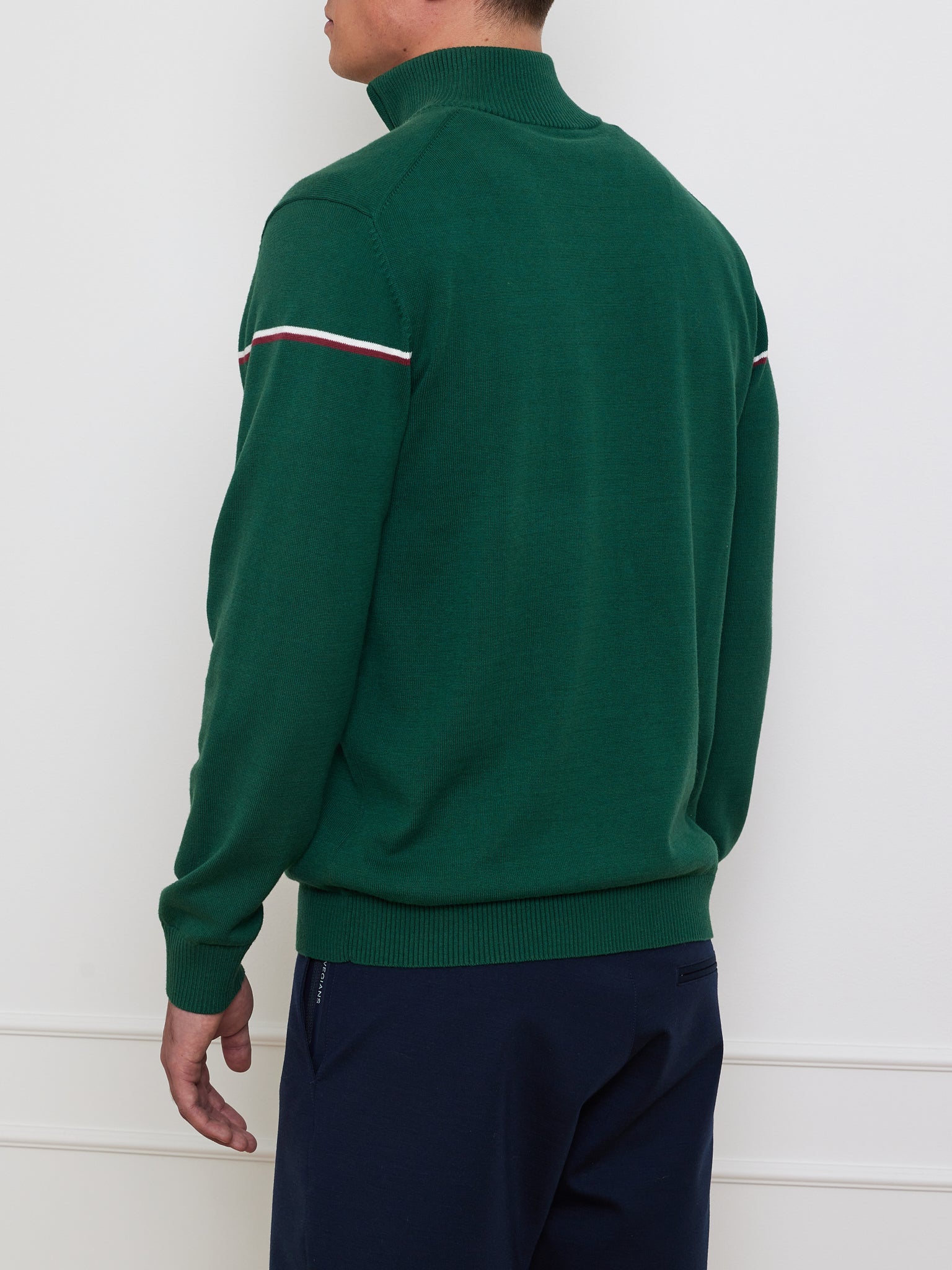 Against a light wall, a person exudes a vintage athletic vibe wearing the "Grand Slam Sweater Men" by We Norwegians. The green long-sleeve sweater features white and red trim with a high collar for an après sport look, complemented by navy pants.