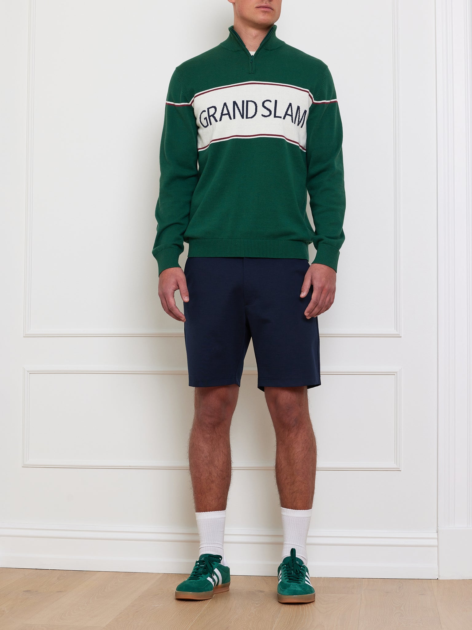 A person stands against a white wall wearing the We Norwegians Grand Slam Sweater Men in green, made from a soft cotton-merino blend, perfect as an après sport layer. Paired with navy shorts, white socks, and green sneakers for vintage athletic vibes.