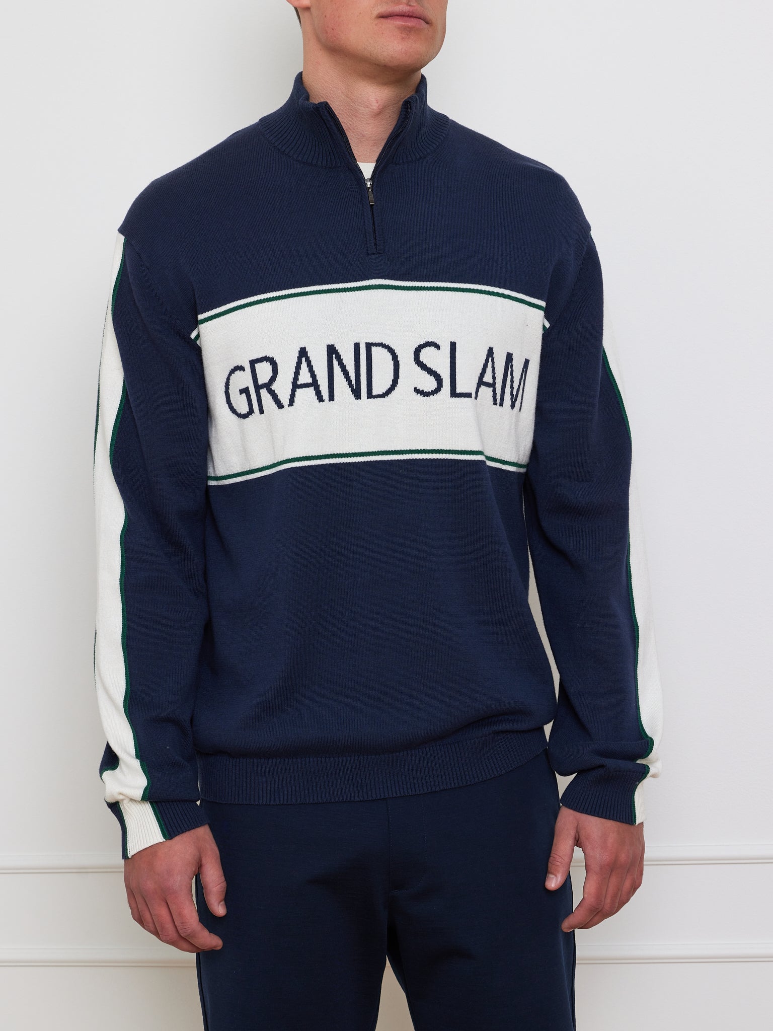 Wearing the "Grand Slam Sweater Men" by We Norwegians, featuring a navy blue zip-up with "GRAND SLAM" on a vintage-inspired white panel, this person pairs it with navy pants. Stylish white sections on the sleeves enhance the classic après-sport look against a plain white wall background.