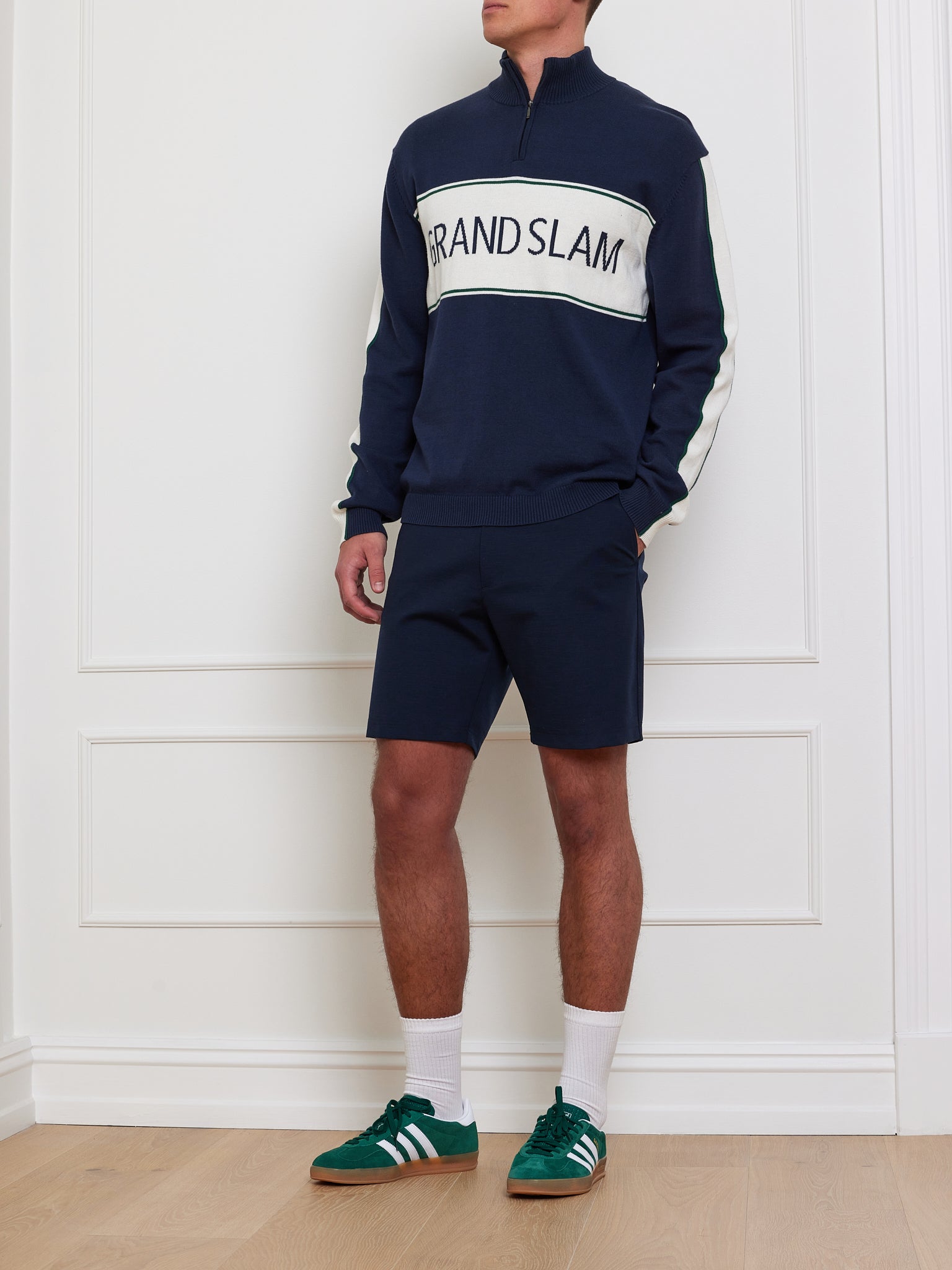 A person stands in a room wearing a "Grand Slam Sweater Men" by We Norwegians, navy shorts, white socks, and green sneakers with white stripes. The background features white paneled walls and a wooden floor.
