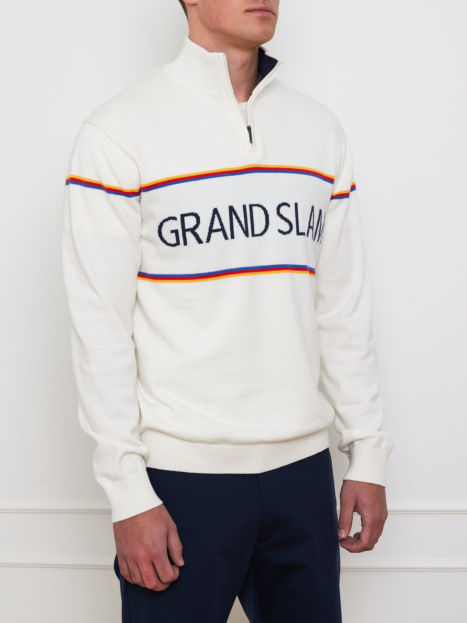 A person rocks a vintage look in the "Grand Slam Sweater Men" by We Norwegians, featuring a half-zip collar with red, yellow, and blue stripes. Made from a cotton-merino blend for an après-sport vibe, it pairs effortlessly with dark pants against a white wall.