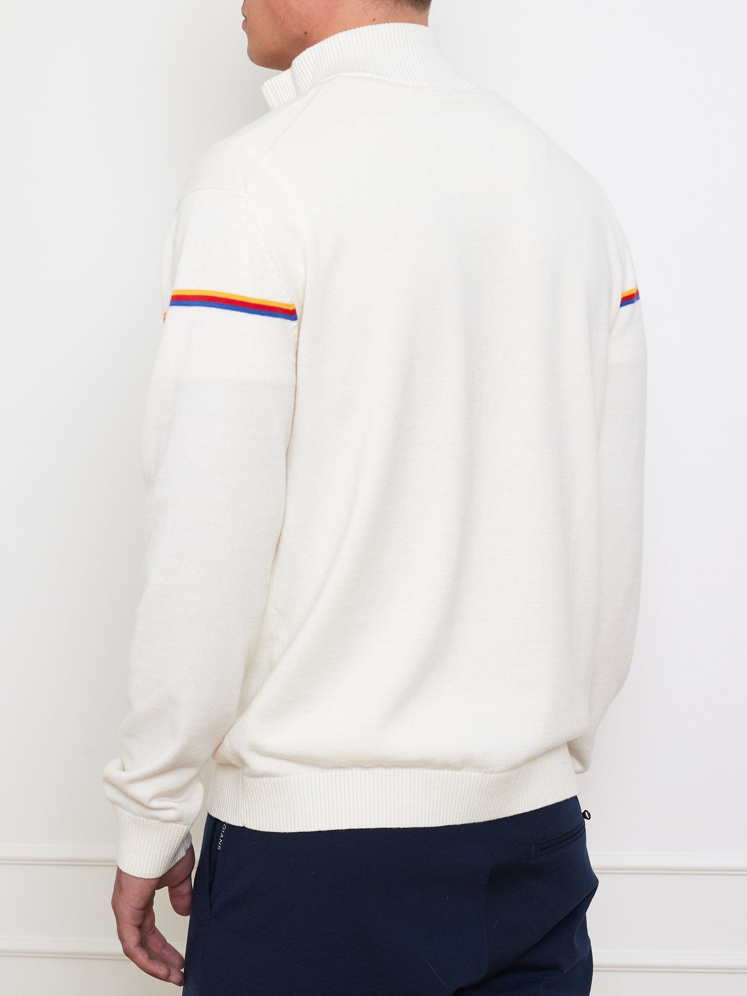 A man, facing away against an off-white wall, wears the We Norwegians Grand Slam Sweater Men in a white cotton-merino blend with rainbow sleeve stripes and a vintage athletic style ribbed collar and cuffs, paired with dark pants.