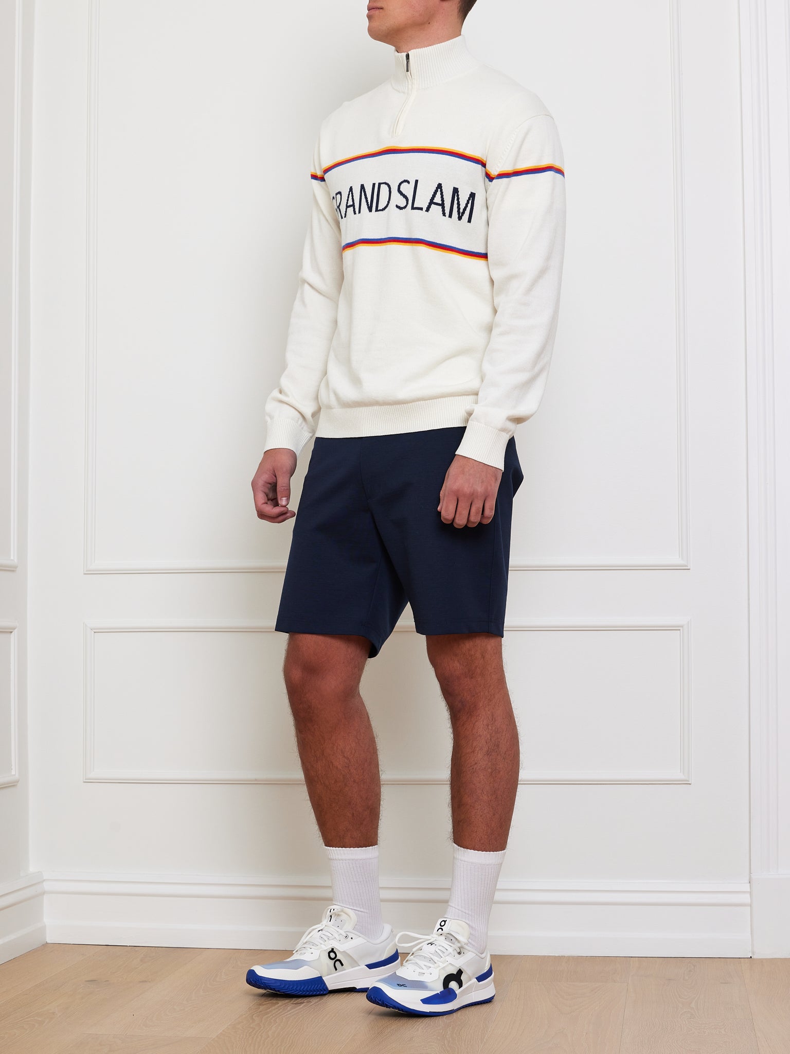 Wearing a Grand Slam Sweater Men by We Norwegians, styled with navy shorts, white socks, and sneakers with blue accents, the individual leans against a sleek white wall. This vintage athletic look is ideal for an après sport vibe.