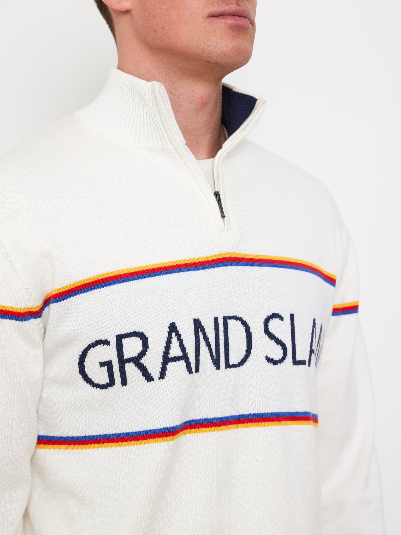 A person models vintage athletic wear in a white Grand Slam Sweater by We Norwegians, featuring a quarter-zip collar, horizontal stripes in blue, orange, and red, and "GRAND SLAM" across the chest against a plain white backdrop.