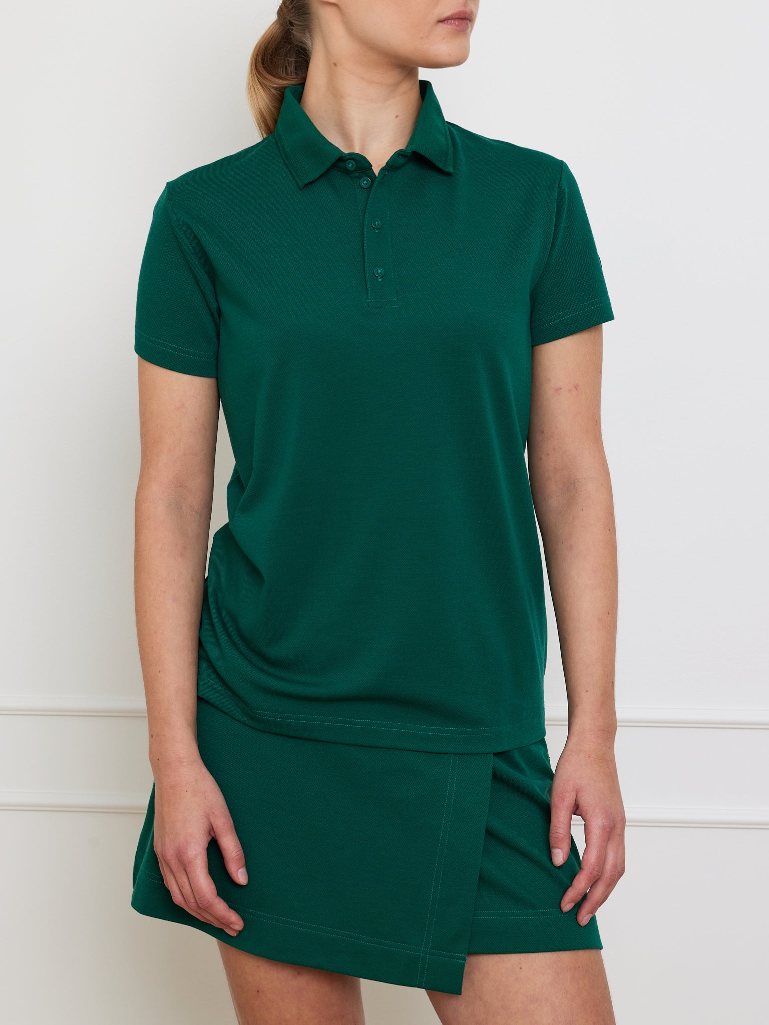 A woman in a We Norwegians Medalist Piqué Polo Women and matching green skirt stands before a white wall. Her hair is pulled back, and her arms rest at her sides.