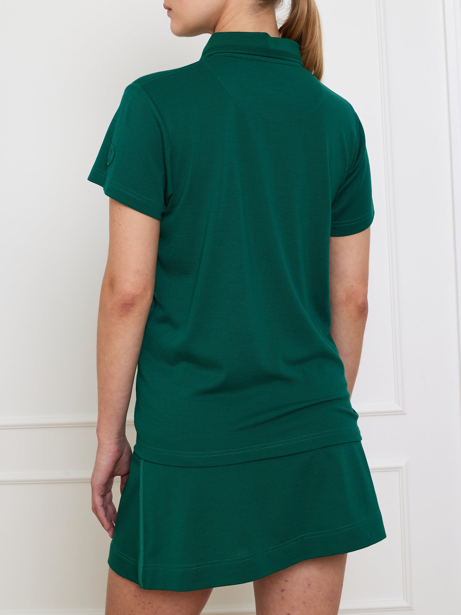 With light brown hair in a ponytail, a person wears a dark green Medalist Piqué Polo by We Norwegians and a matching skirt, standing in front of a white panel wall, facing away from the camera.