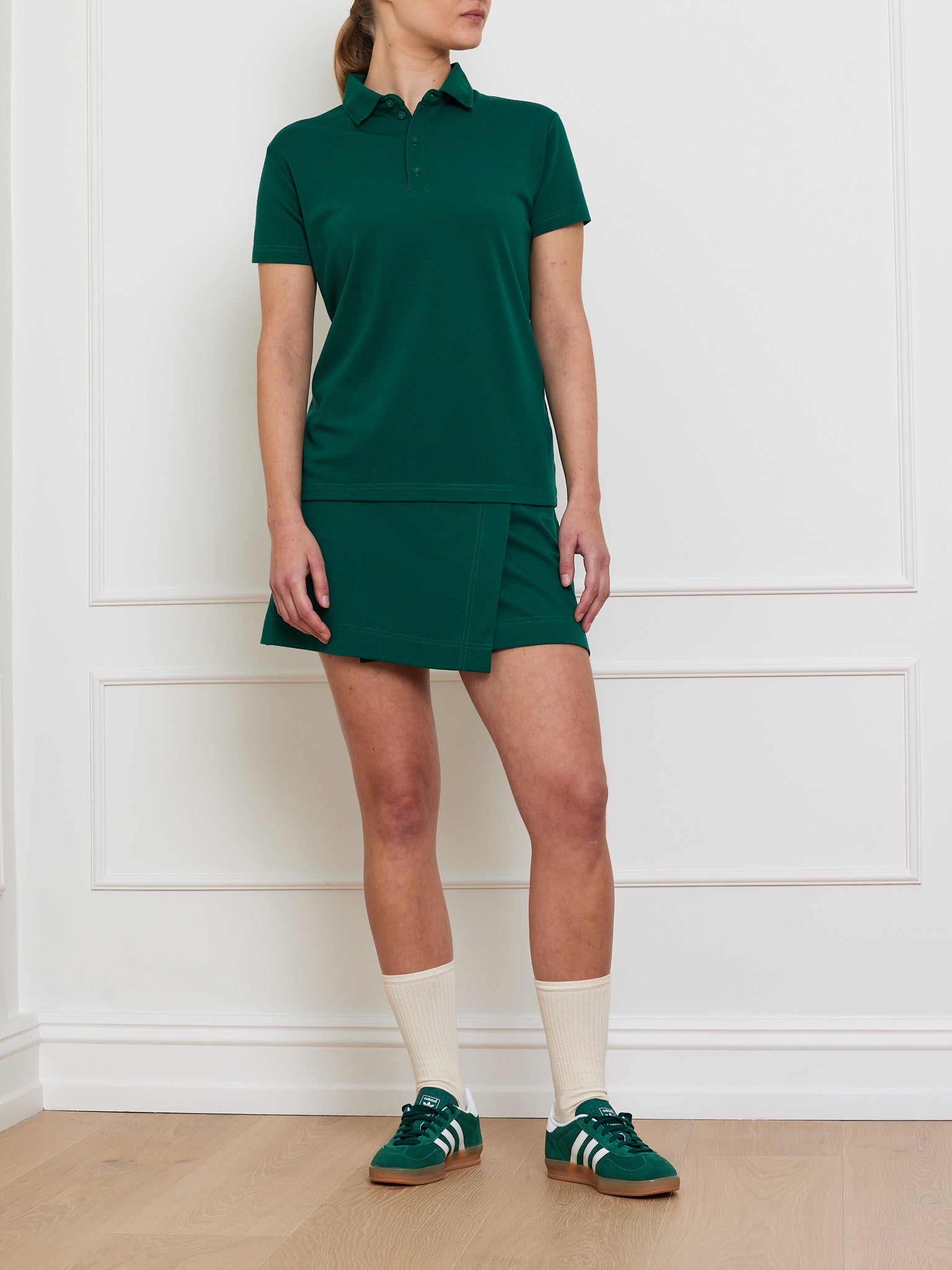 A We Norwegians Medalist Piqué Polo in green and a matching skirt are worn by a person against a white wall. They have on green sneakers with white stripes, cream mid-calf socks, and the light wood floor suggests an indoor setting.