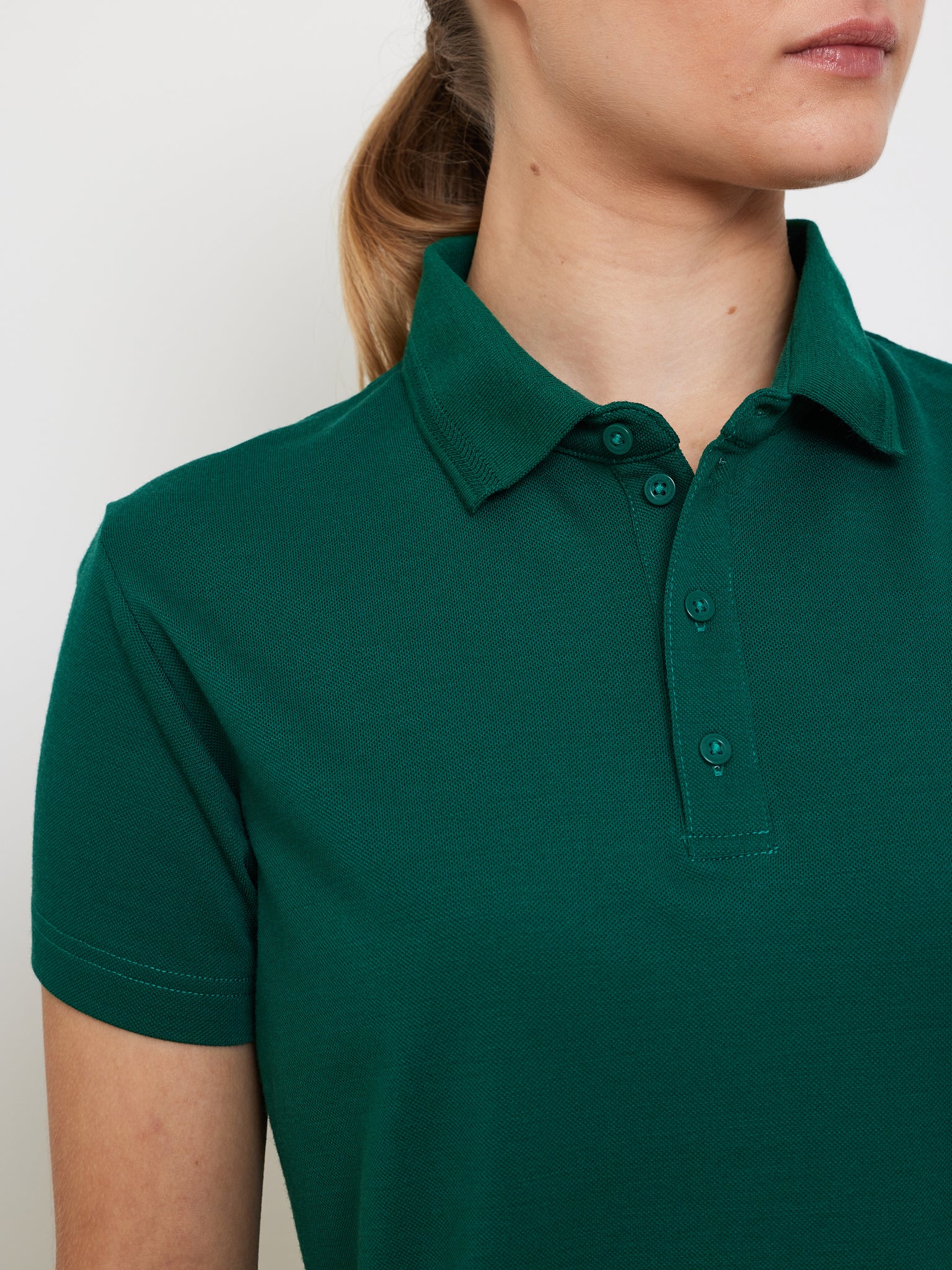 A person in a green We Norwegians Medalist Piqué Polo Women with a buttoned collar and moisture-wicking fabric stands against a plain background, hair tied back.