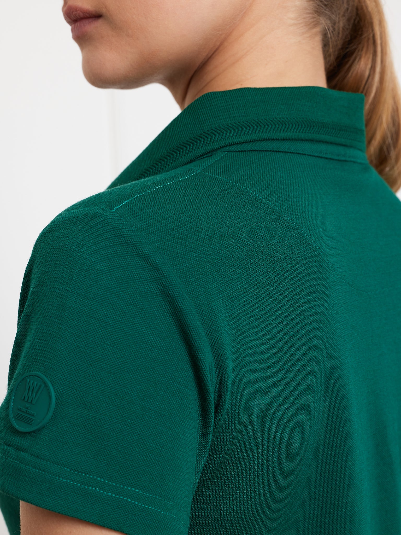 Close-up of a person wearing the Medalist Piqué Polo Women by We Norwegians, highlighting the green, moisture-wicking textured fabric on the shoulder and collar. The sleeve features a circular logo with embossed text and graphics, while the person's hair is tied back.