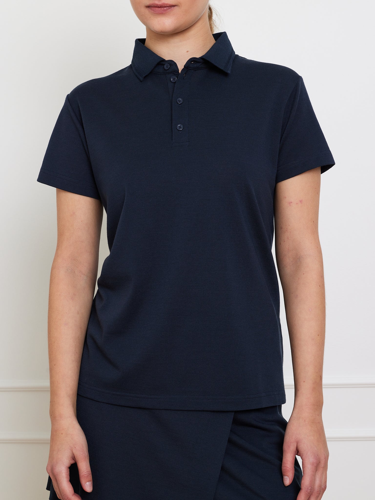 Someone is wearing the We Norwegians Medalist Piqué Polo Women in navy blue, featuring a collar and buttoned placket. They stand with arms relaxed by their sides against a plain white wall, with their face not visible.