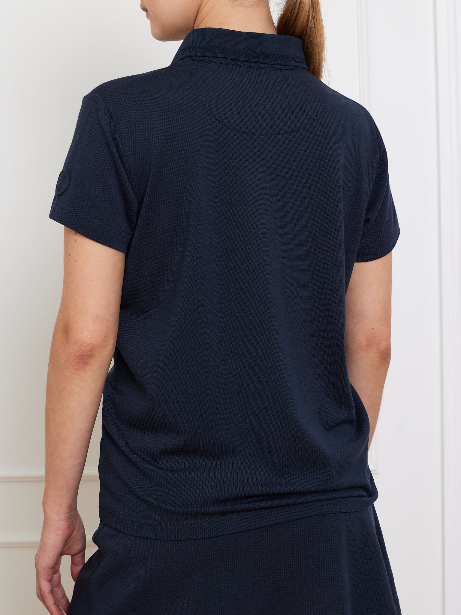 A person with light skin and long hair tied back wears a navy Medalist Piqué Polo Women by We Norwegians, paired with a matching skirt, standing against a subtly paneled white wall.