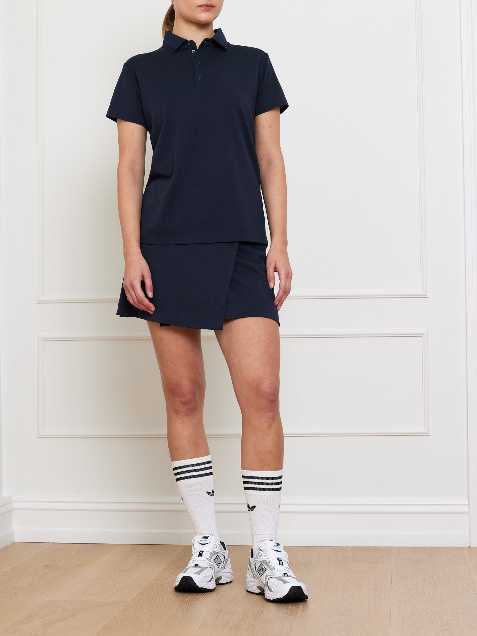 A person is elegantly dressed indoors, wearing a We Norwegians Medalist Piqué Polo for women with a matching skirt. They finish the look with white socks featuring black stripes and white sneakers against a white paneled wall and wooden floor, showcasing casual sophistication.