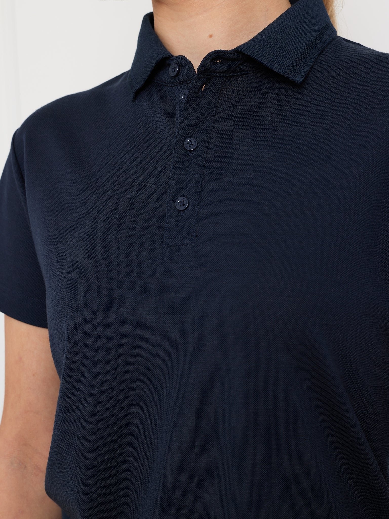 A person is wearing the Medalist Piqué Polo Women by We Norwegians. This dark navy blue polo features short sleeves, a collar with three buttons, and moisture-wicking fabric for all-day comfort against a plain, light-colored background.