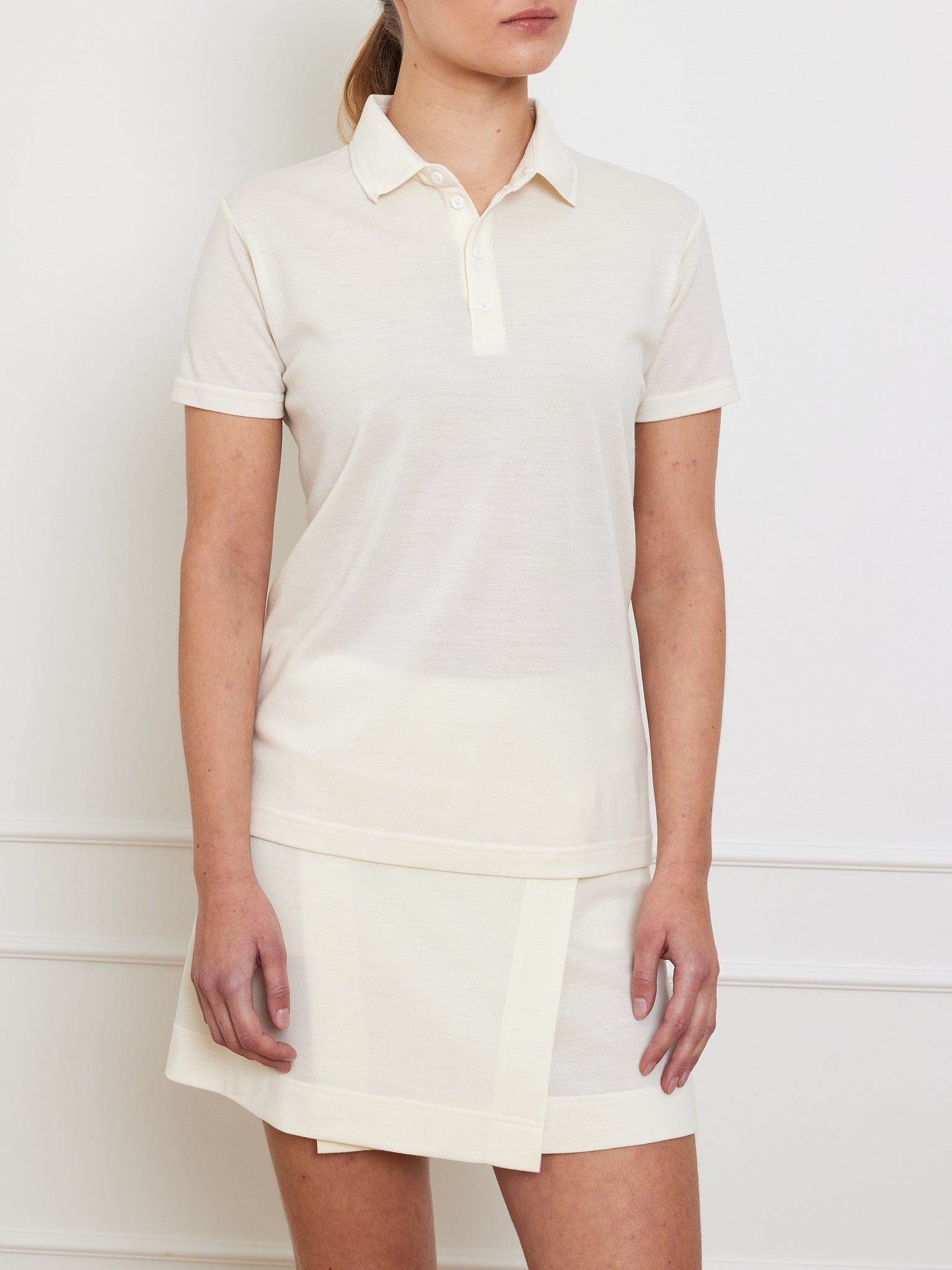 A person is seen against a plain white wall dressed in the simple and elegant Medalist Piqué Polo Women's shirt and matching skirt by We Norwegians. The cream-colored outfit features short sleeves and a classic collared design, but the wearer's face isn't visible.