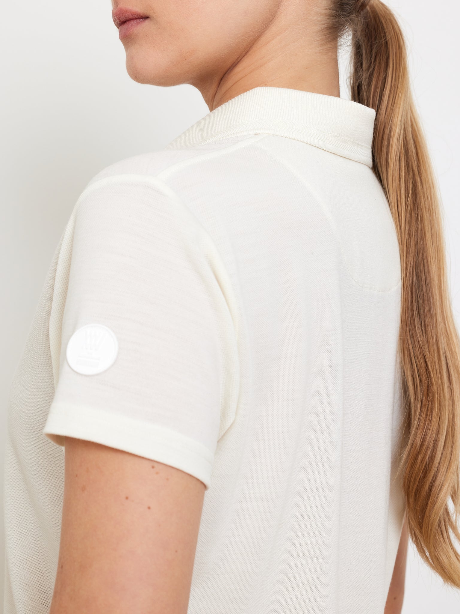 Someone with long hair in a ponytail is wearing a "Medalist Piqué Polo Women" by We Norwegians. The polo, which is off-white and moisture-wicking, has a circular white patch on the sleeve. The person's face remains unseen against the plain white backdrop.