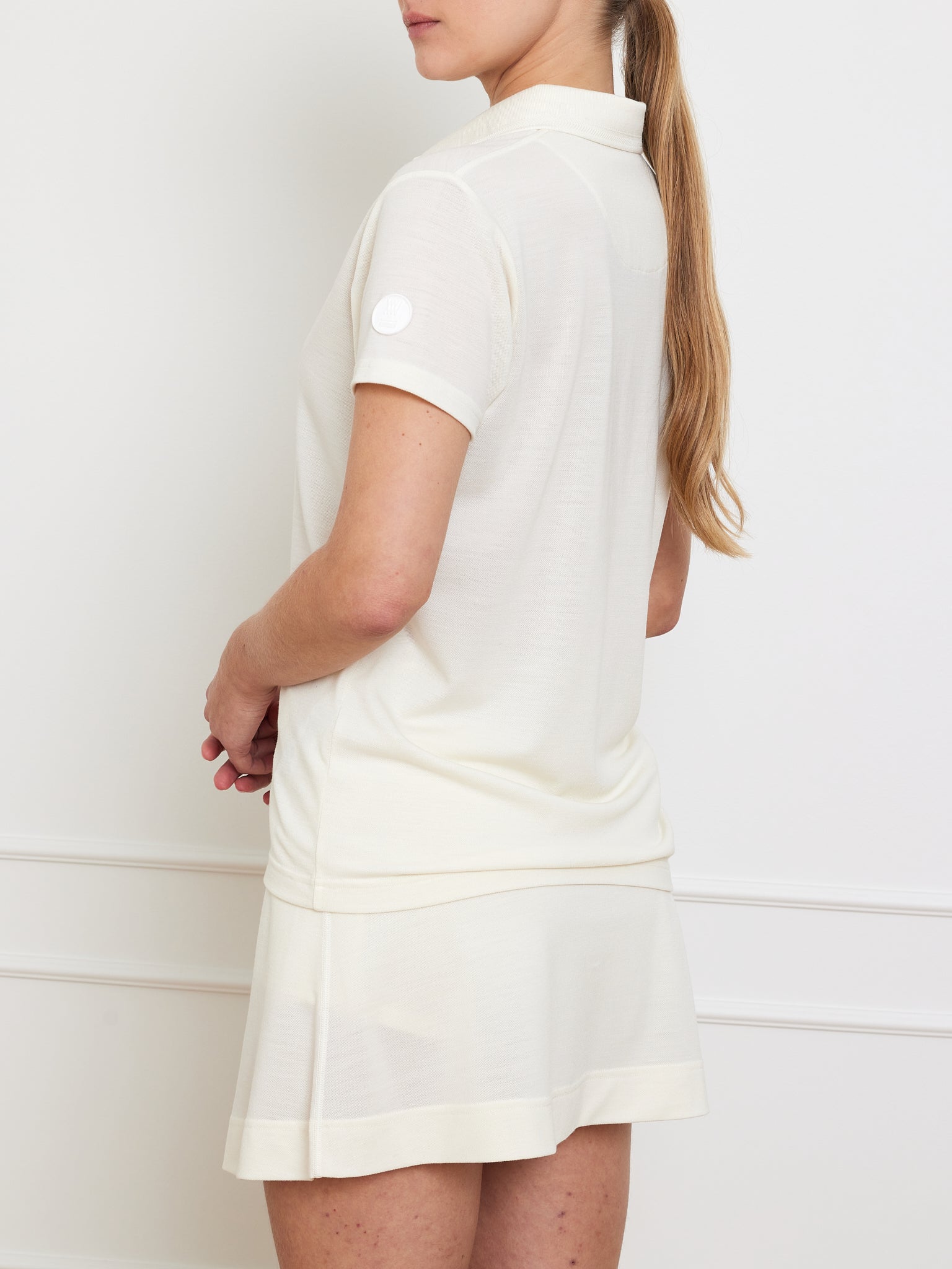 A person with long hair stands sideways against a plain white background, wearing a light Medalist Piqué Polo Women and matching skirt by We Norwegians, with their arms relaxed at their sides.