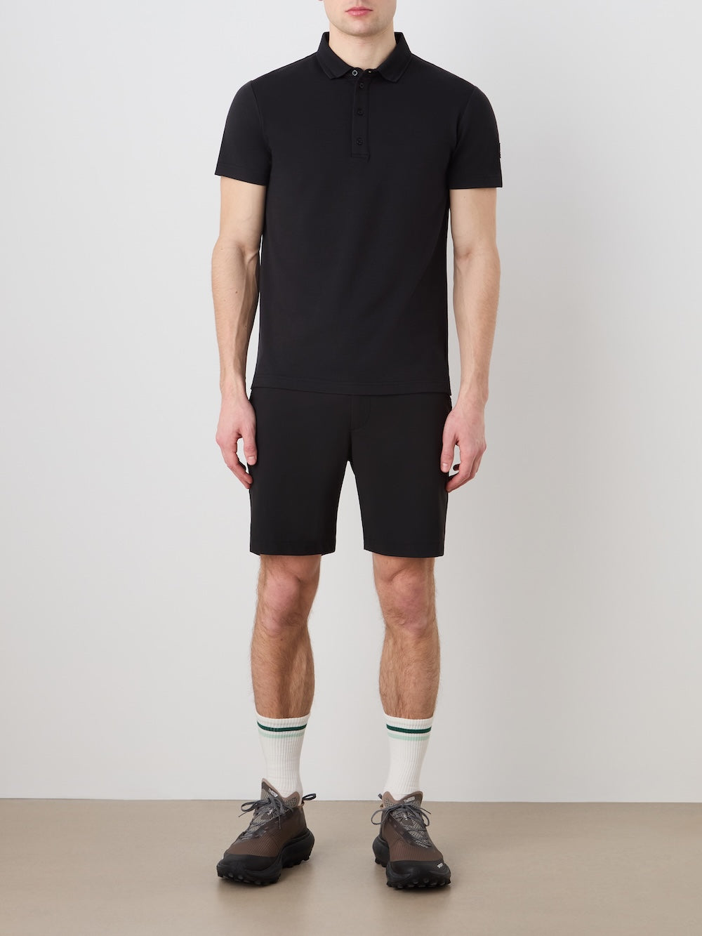 A man stands against a light wall wearing the Medalist Piqué Polo Men by We Norwegians, paired with black shorts, white socks with green stripes, and black athletic shoes. His arms hang by his sides.