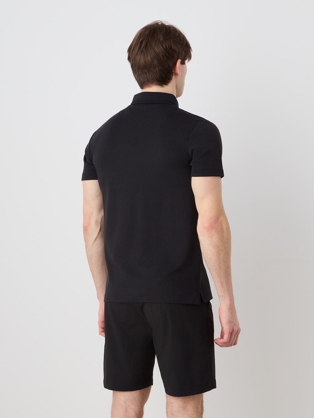 A man with light skin and brown hair faces away from the camera, wearing a black We Norwegians Medalist Piqué Polo Men and black shorts against a plain light background.