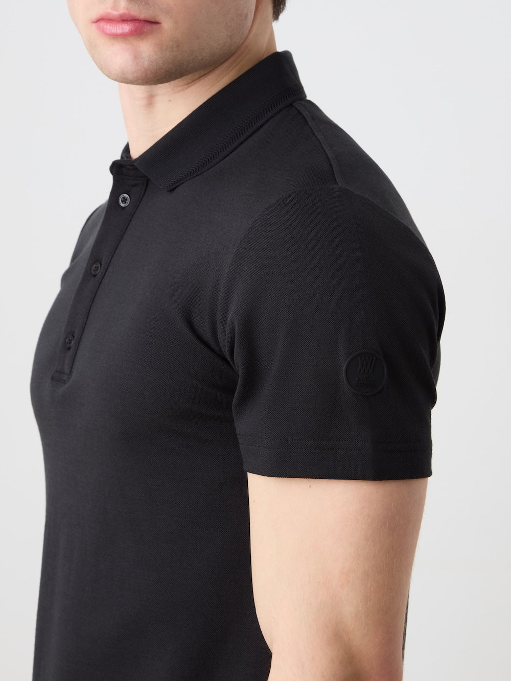 A man wearing the We Norwegians Medalist Piqué Polo Men, a fitted black moisture-wicking shirt with a subtle logo on the left sleeve, is shown from the chest up against a plain light background.