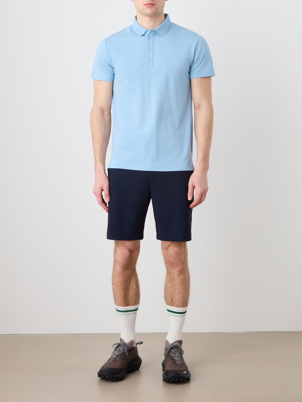 A person stands against a plain background wearing the We Norwegians Medalist Piqué Polo Men in light blue, paired with navy shorts, white socks featuring green stripes, and gray hiking shoes. The individual's face is not visible.