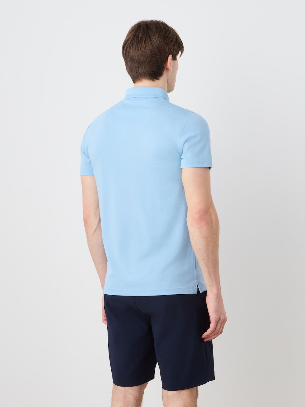 A person with short brown hair faces away from the camera, wearing the We Norwegians Medalist Piqué Polo Men in light blue and dark navy shorts against a plain white background.