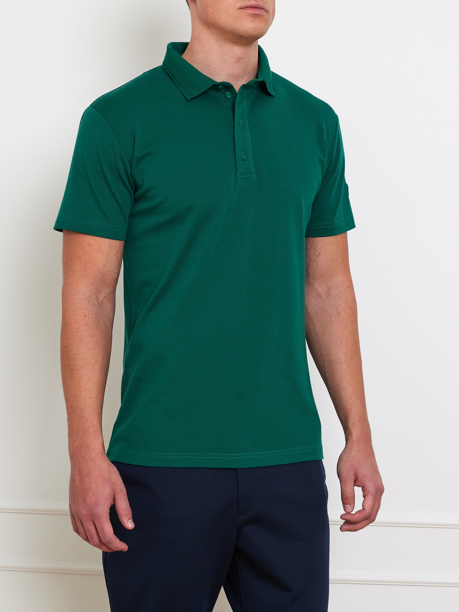 A man is wearing a We Norwegians Medalist Piqué Polo Men in dark green, crafted from CoolMax fabric with short sleeves, posing against a white backdrop. Only his upper body is shown.