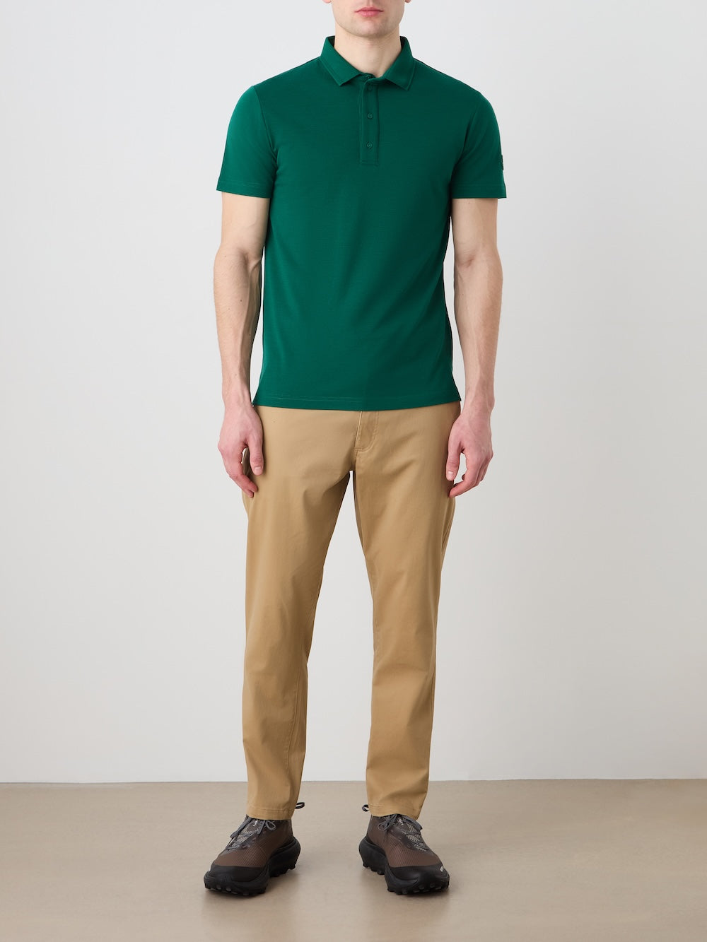 A man stands against a plain background, wearing the We Norwegians Medalist Piqué Polo Men in green, beige trousers, and dark brown shoes. His arms are relaxed at his sides and his expression is neutral.