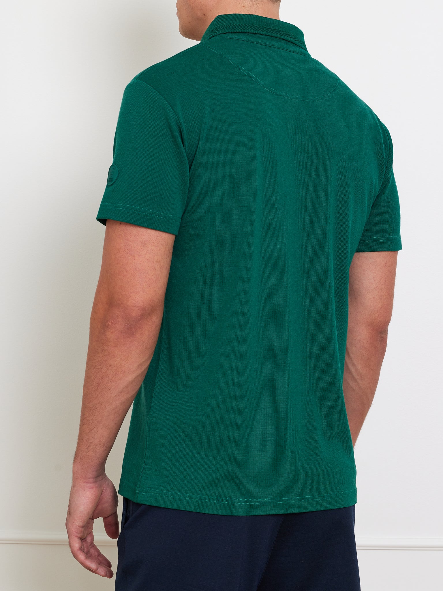 Rear view of a person wearing the Medalist Piqué Polo Men by We Norwegians in dark green, paired with dark pants against a plain white wall.