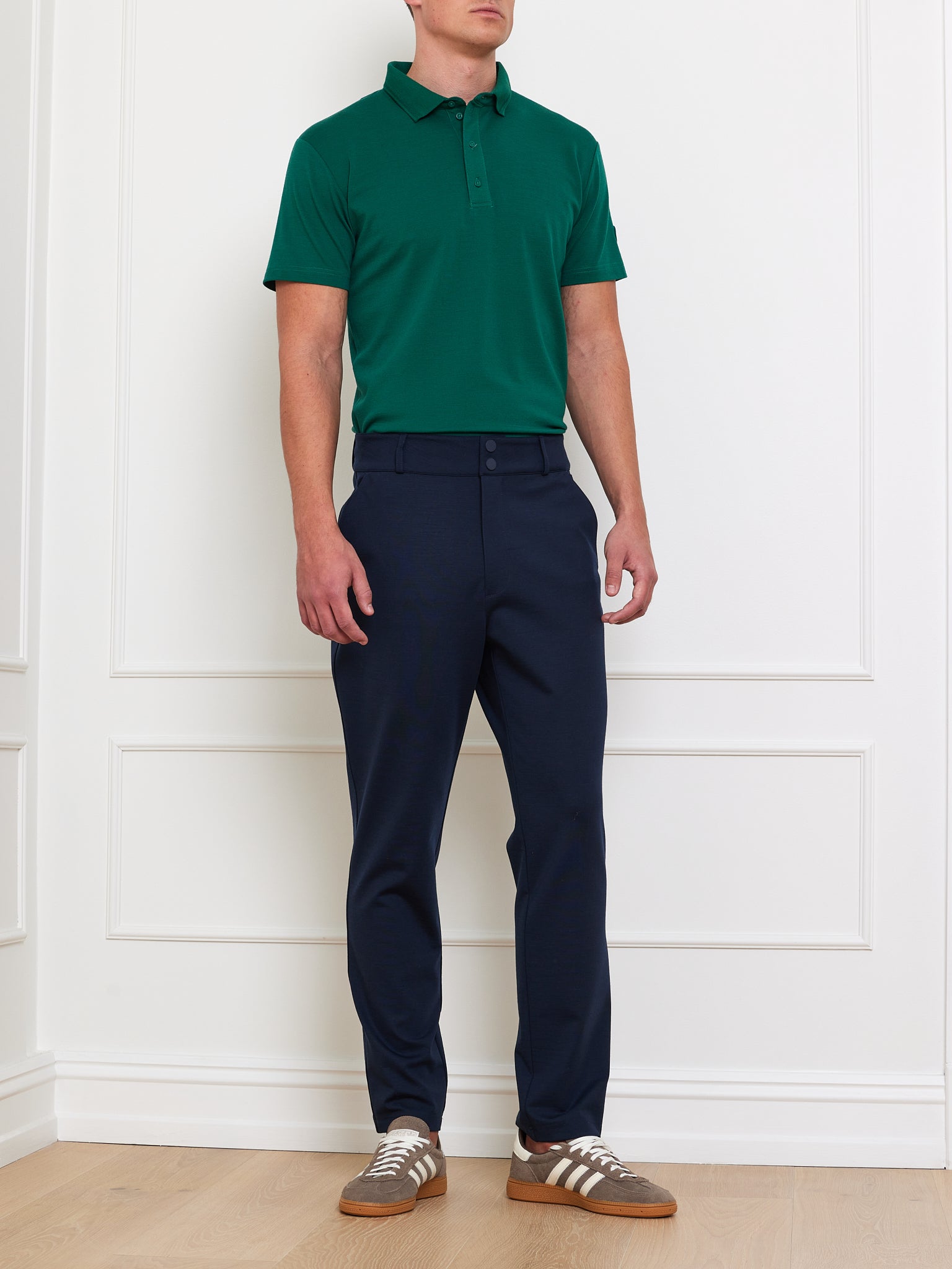 A man in a We Norwegians Medalist Piqué Polo Men stands against a white paneled wall, wearing navy blue pants and brown shoes with white stripes, posing casually with arms by his sides.