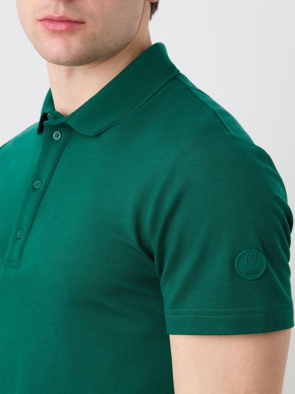 A person wearing the We Norwegians Medalist Piqué Polo Men in green, a short-sleeve moisture-wicking shirt with a subtle embroidered sleeve logo, shown from shoulders to mid-chest against a plain light background.