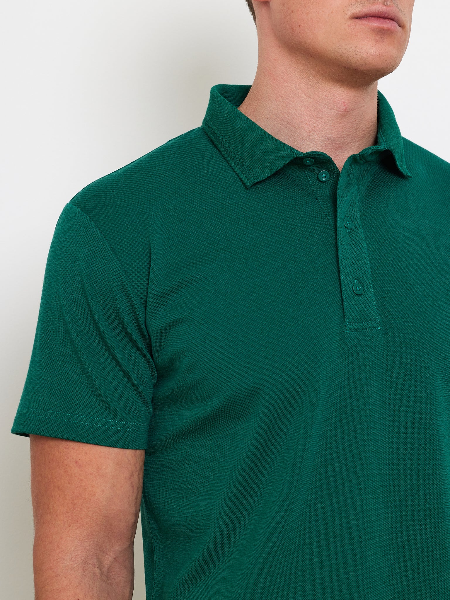 A person in a "We Norwegians" Medalist Piqué Polo, made with CoolMax fabric, stands against a white background. The green shirt features a collar and buttons. The image focuses on the upper torso, subtly obscuring the face for an anonymous yet stylish appeal.