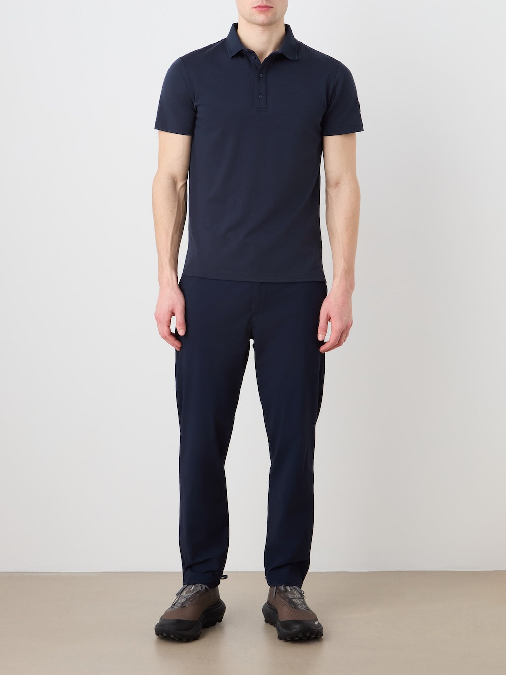 A man stands against a plain light background wearing the We Norwegians Medalist Piqué Polo Men in navy, matching navy pants, and dark athletic shoes. His arms are relaxed at his sides and his face is partially out of frame.