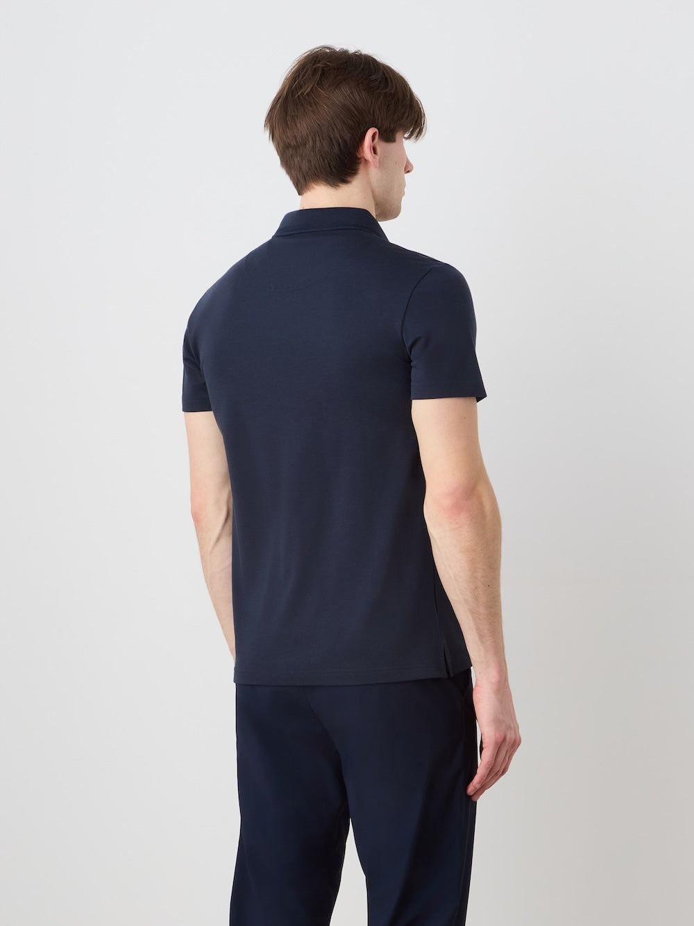 A person with short brown hair stands facing away from the camera, wearing a We Norwegians Medalist Piqué Polo Men in dark navy and matching pants, set against a plain light background.