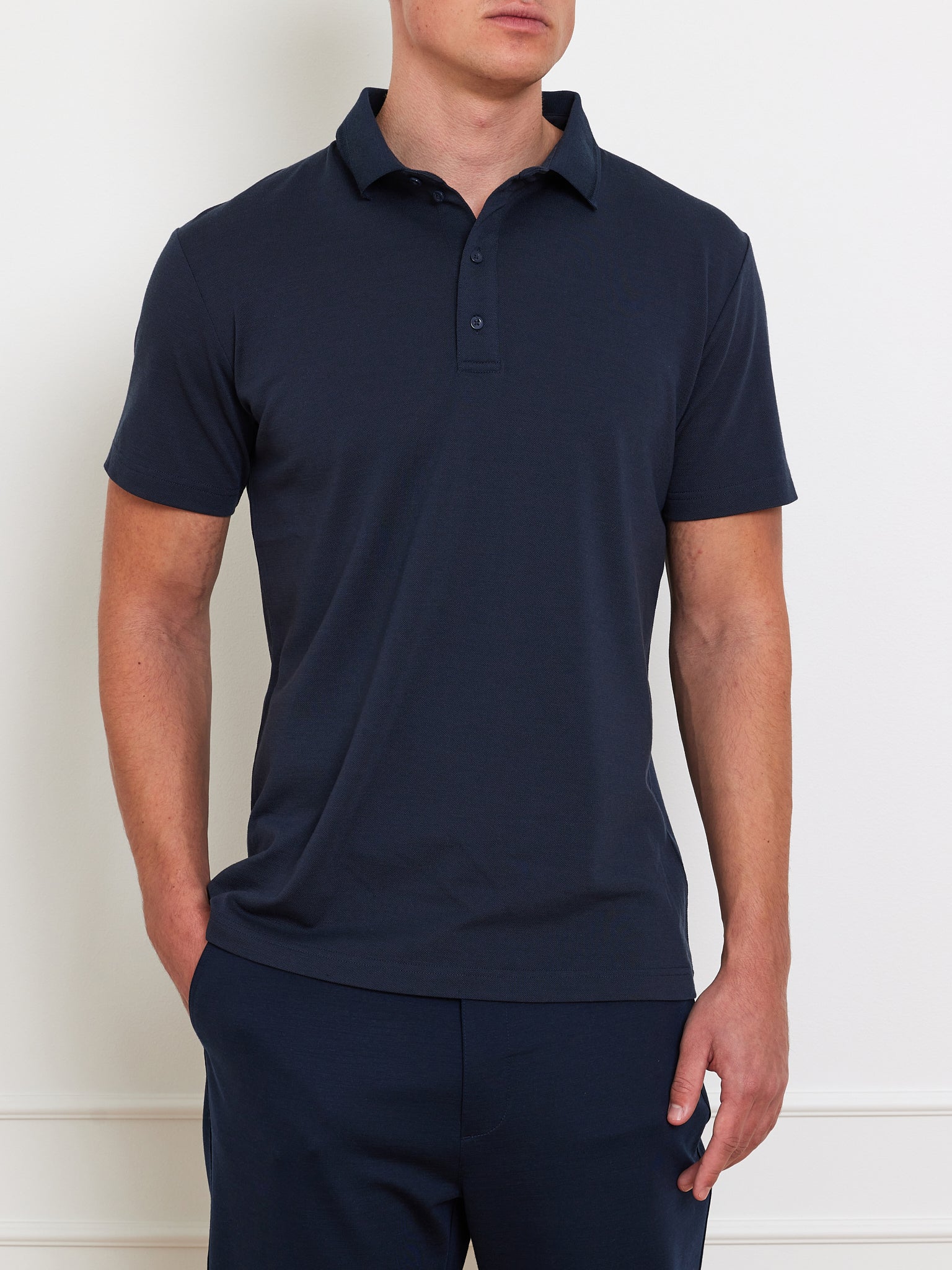 A person wearing a dark navy Medalist Piqué Polo by We Norwegians and matching pants stands against a light background, one hand in their pocket. The polo features a collar and three buttons, ideal for keeping cool and comfortable.