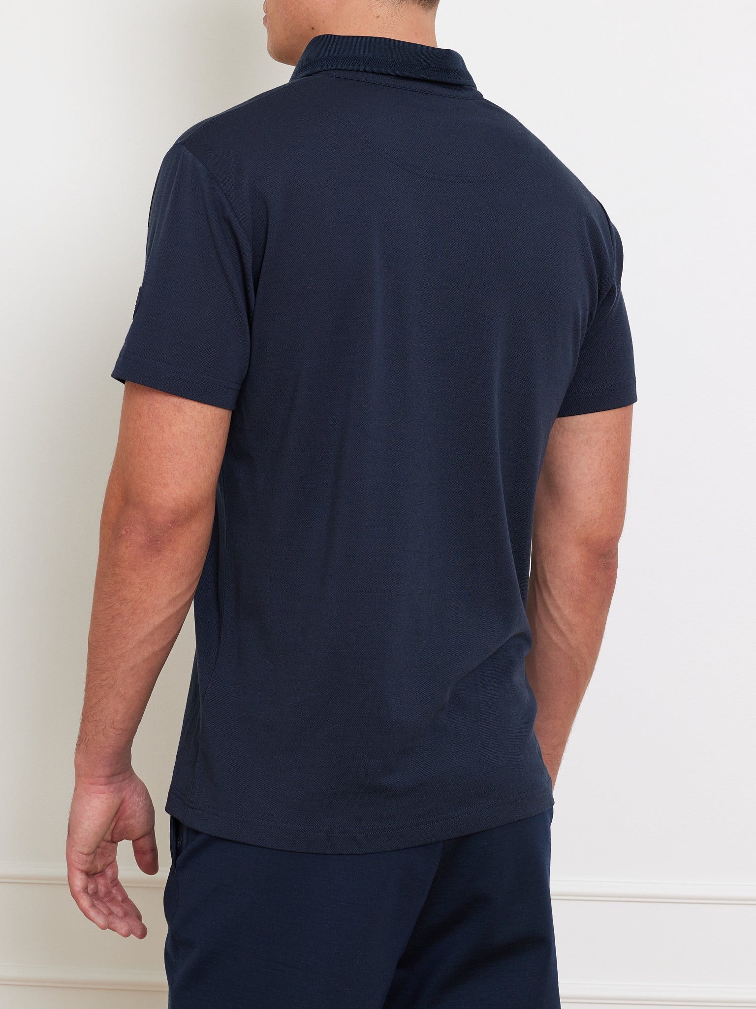 A person in a navy blue We Norwegians Medalist Piqué Polo stands with their back to the camera, complemented by matching pants in a minimalist indoor space against a light-colored wall.