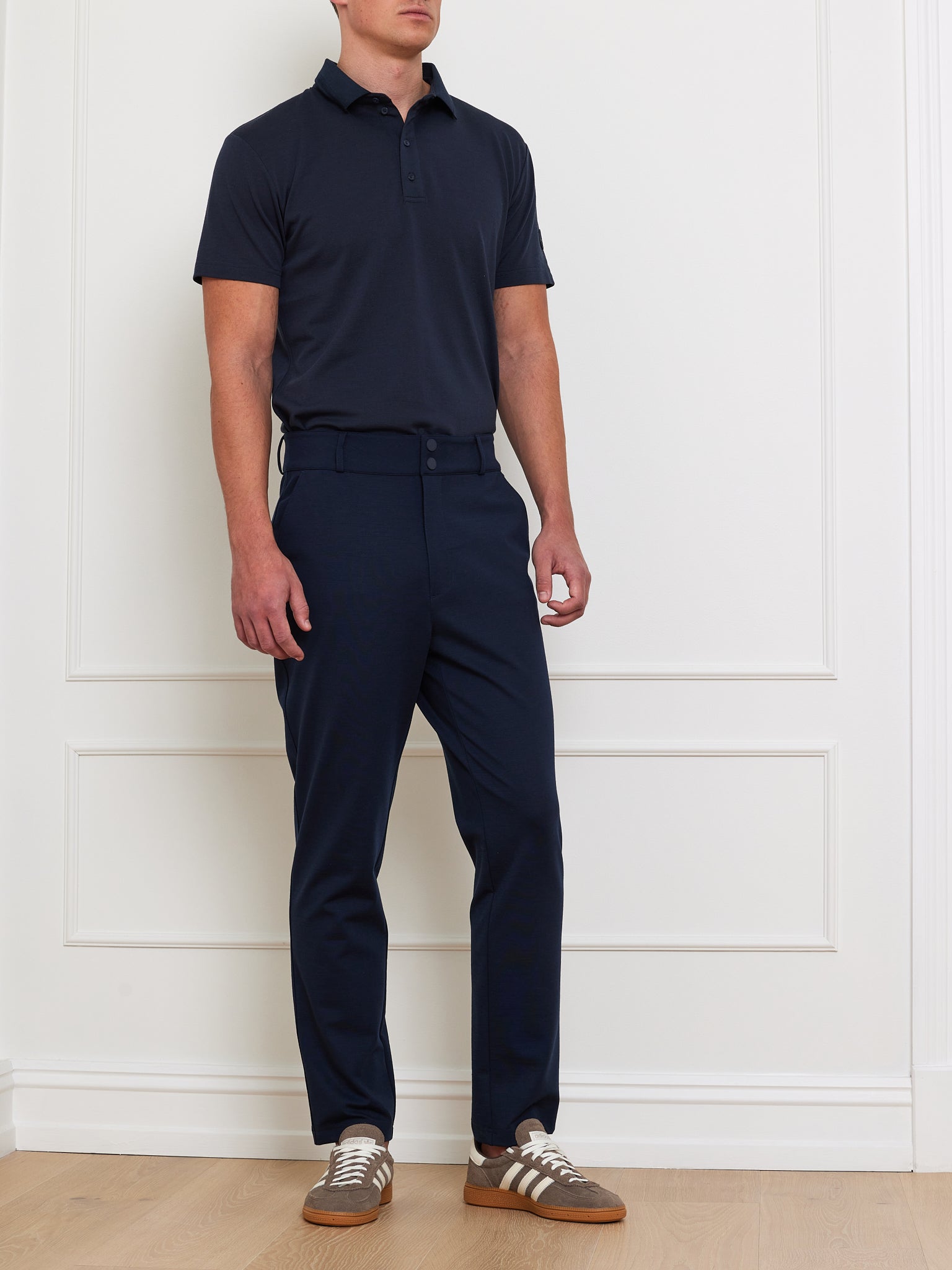 A person stands against a white wall in a dark We Norwegians Medalist Piqué Polo Men shirt, paired with matching trousers and a belt. Brown shoes with white stripes complete the look, all set against the backdrop of a wooden floor.
