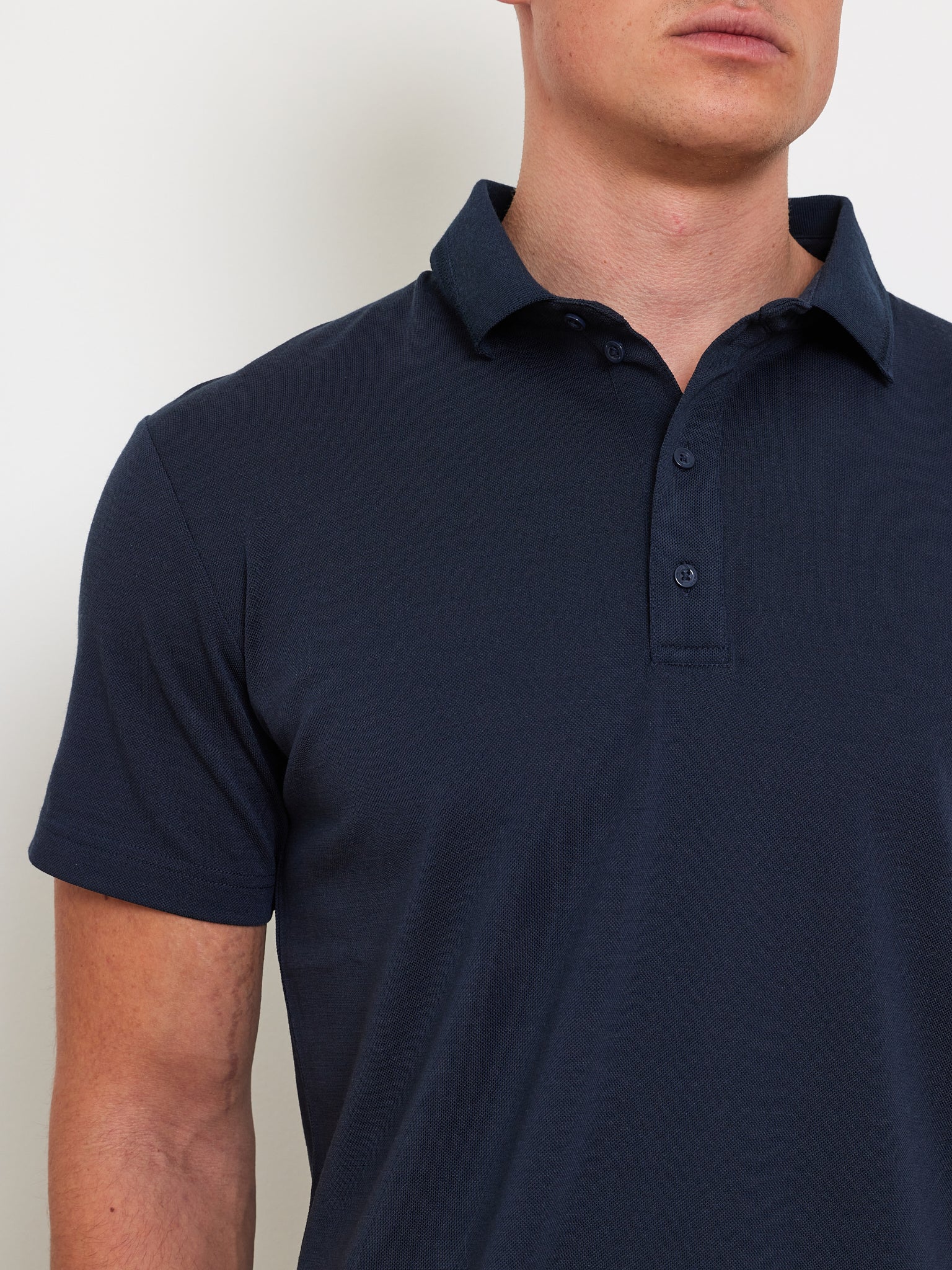 A person wearing the Medalist Piqué Polo Men by We Norwegians, crafted from CoolMax fabric in dark blue, stands against a plain white background. The shirt has a classic collar and three buttons. Only the upper body and part of the face are visible.