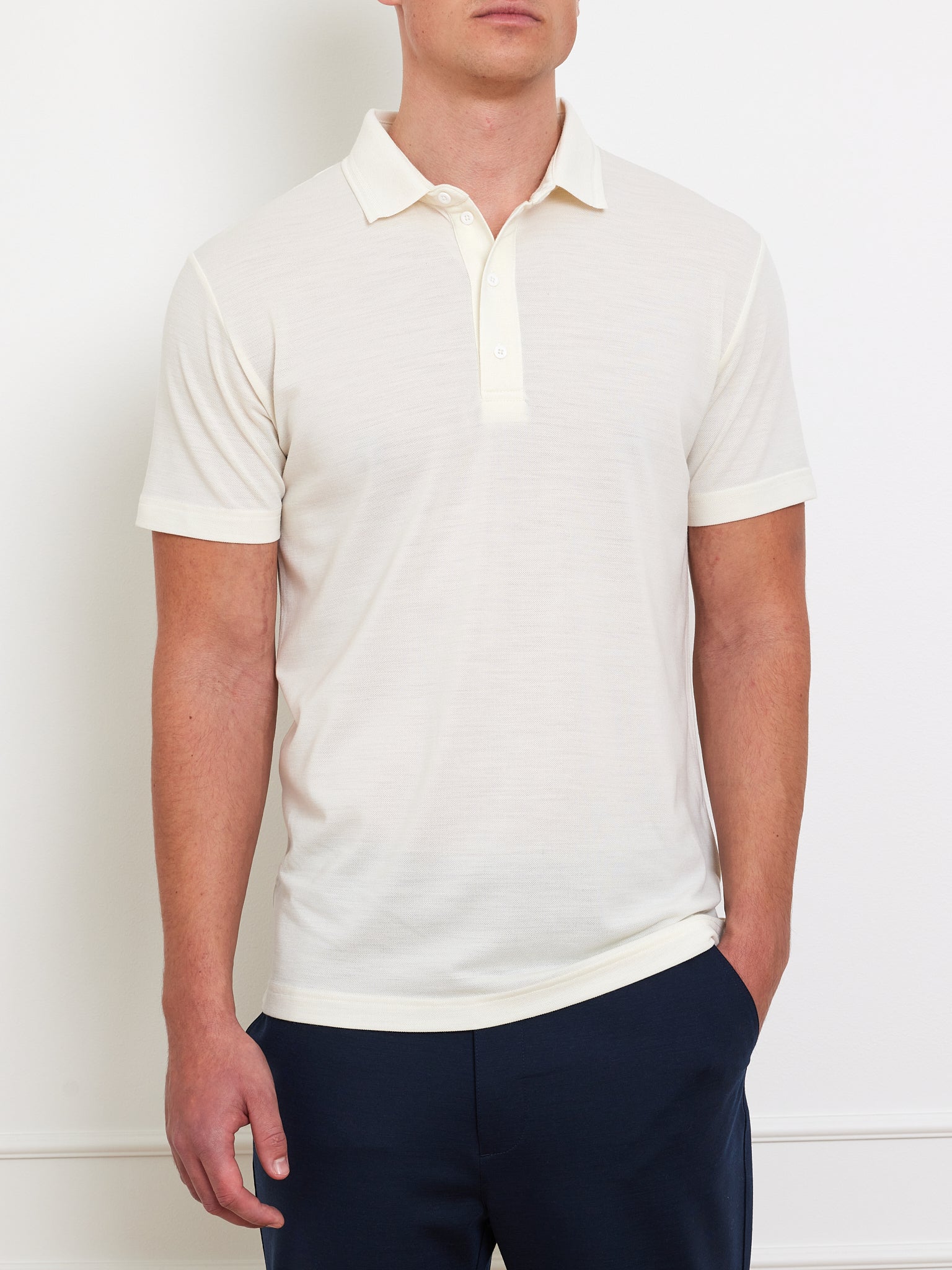 A person wearing a cream Medalist Piqué Polo Men by We Norwegians and dark navy pants poses against a plain white background, left hand in pocket, right arm relaxed at their side.