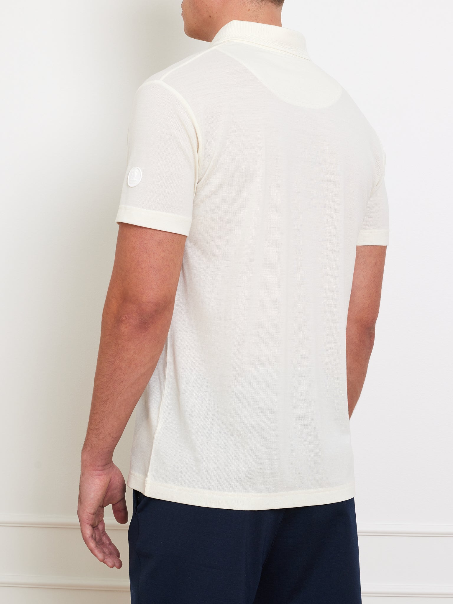 A person in the Medalist Piqué Polo Men by We Norwegians stands facing a white wall. The polo, featuring a left sleeve logo, blends style with innovative fabric technology. They pair it with dark blue pants for a complete look.