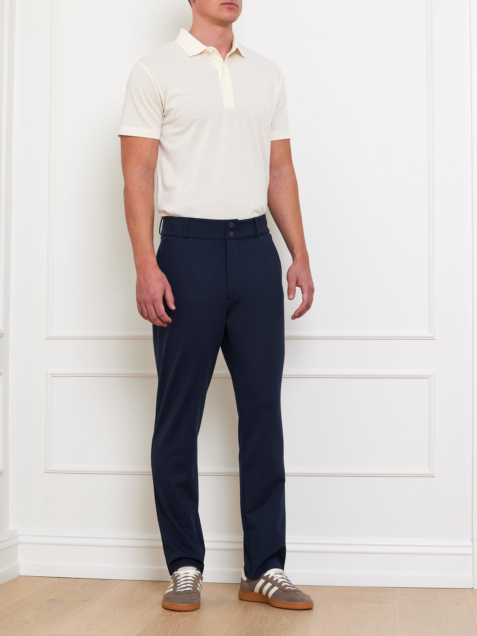 Against a paneled wall, a person wears a cream Medalist Piqué Polo by We Norwegians, dark blue pants, and brown sneakers with white laces on a light wood floor.