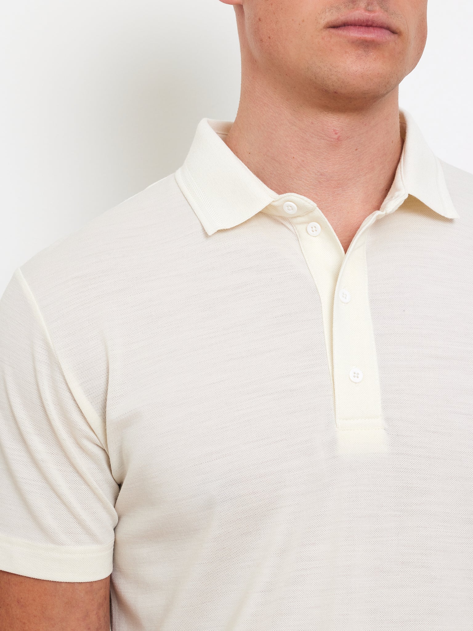 A person wearing a cream Medalist Piqué Polo by We Norwegians stands against a plain background. The shirt, featuring textured fabric and a buttoned collar, provides style and moisture-wicking comfort. Only the torso and part of the face are visible.