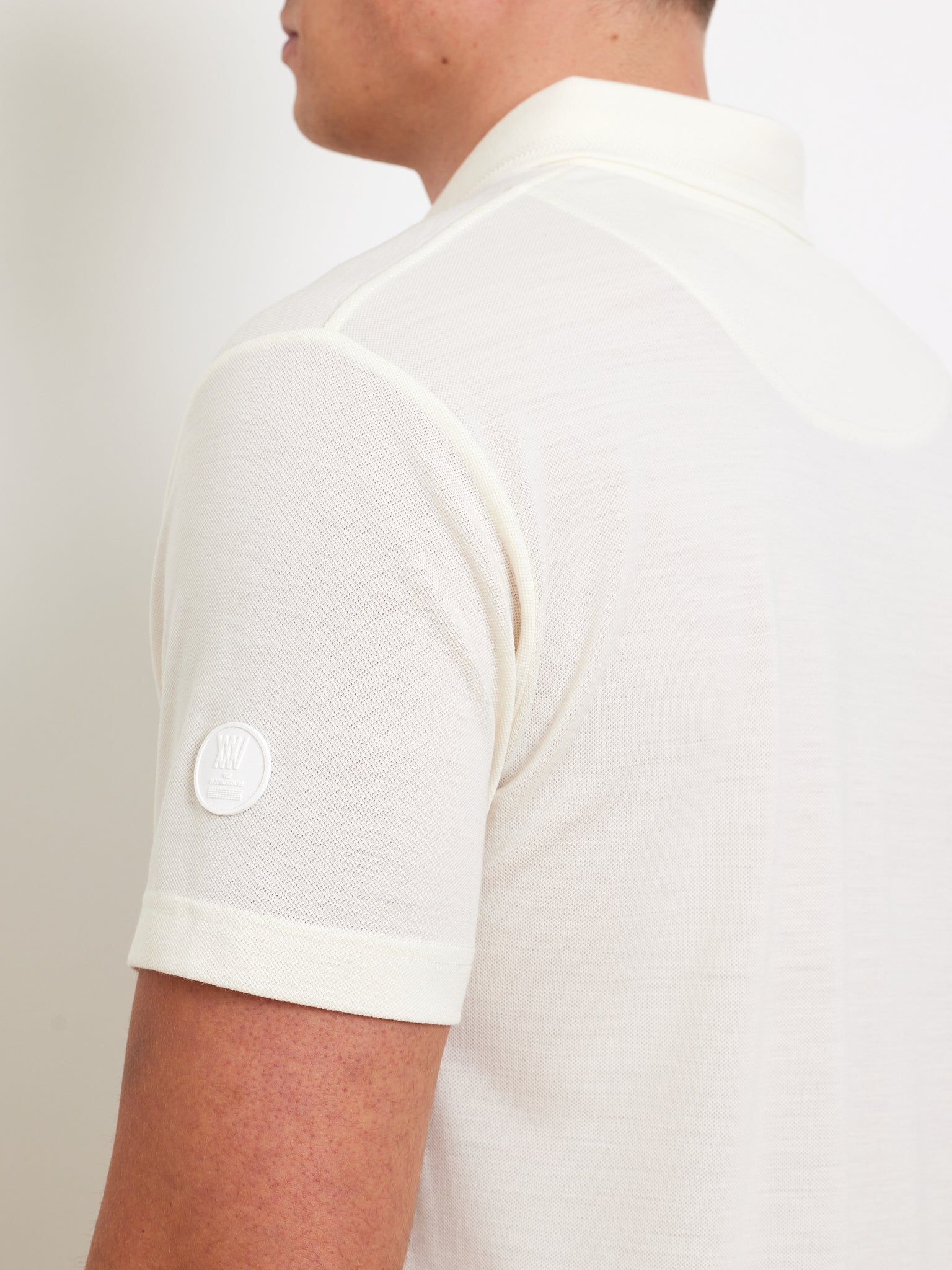 A person wears the Medalist Piqué Polo Men by We Norwegians, a light-colored, short-sleeve shirt made from CoolMax fabric. The side and back reveal a small circular logo on the sleeve. This polo blends style with functionality against a plain white background.