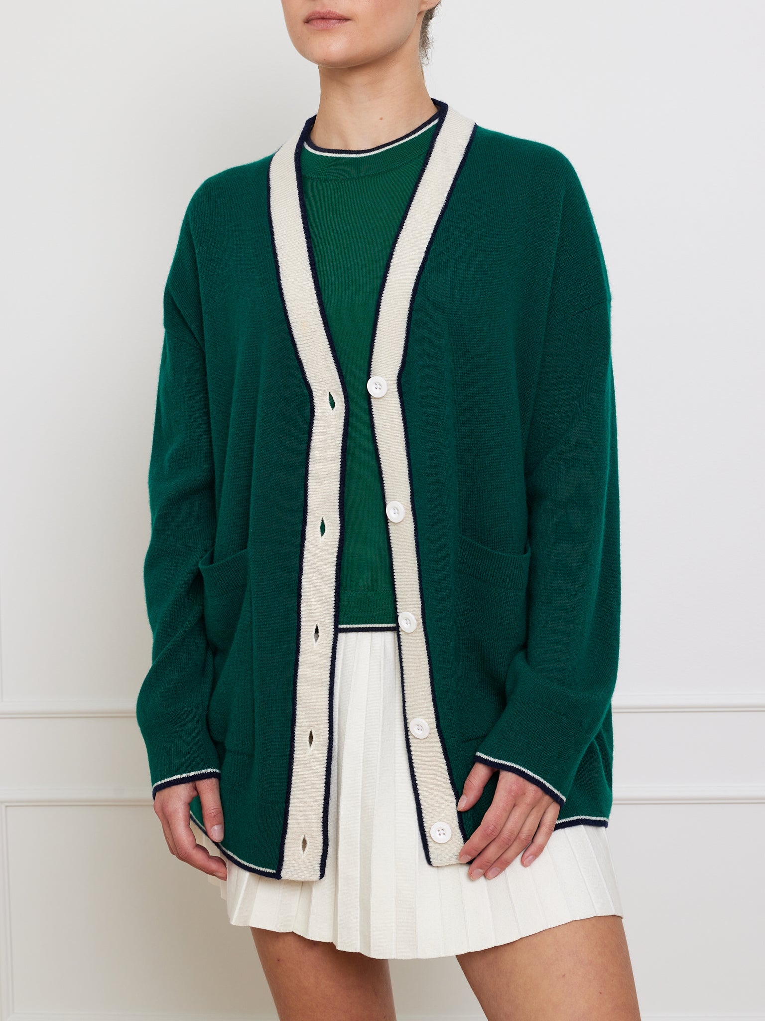 A person is elegantly dressed in the We Norwegians Club Cardigan Women, a luxurious dark green cashmere piece with contrasting light trim and buttons, layered over a matching top. The oversized look is completed with a crisp white pleated skirt. Their head is not visible.