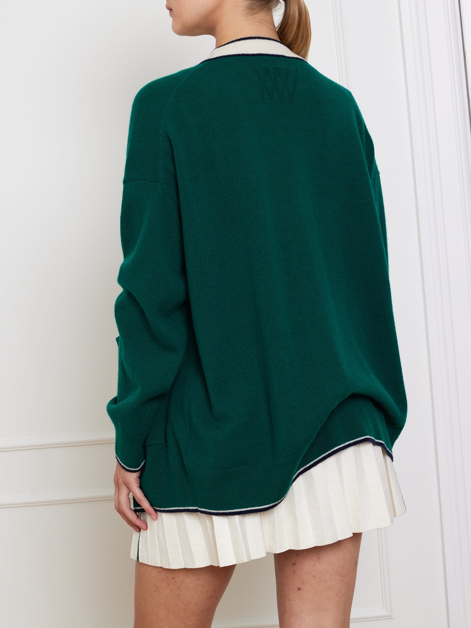 A person wearing an oversized green Club Cardigan Women by We Norwegians, with embroidered details on the back, a white collar, and a white pleated skirt stands facing a white paneled wall.