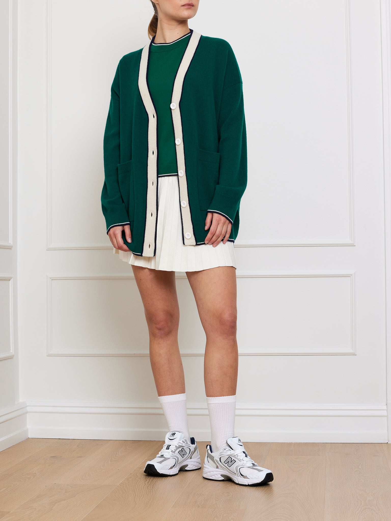 A person exudes elegance and charm, wearing the luxurious We Norwegians Club Cardigan in green with white trim over a green top. Paired with a white pleated skirt, white socks, and sneakers, they stand on a wooden floor against a sleek white paneled wall.