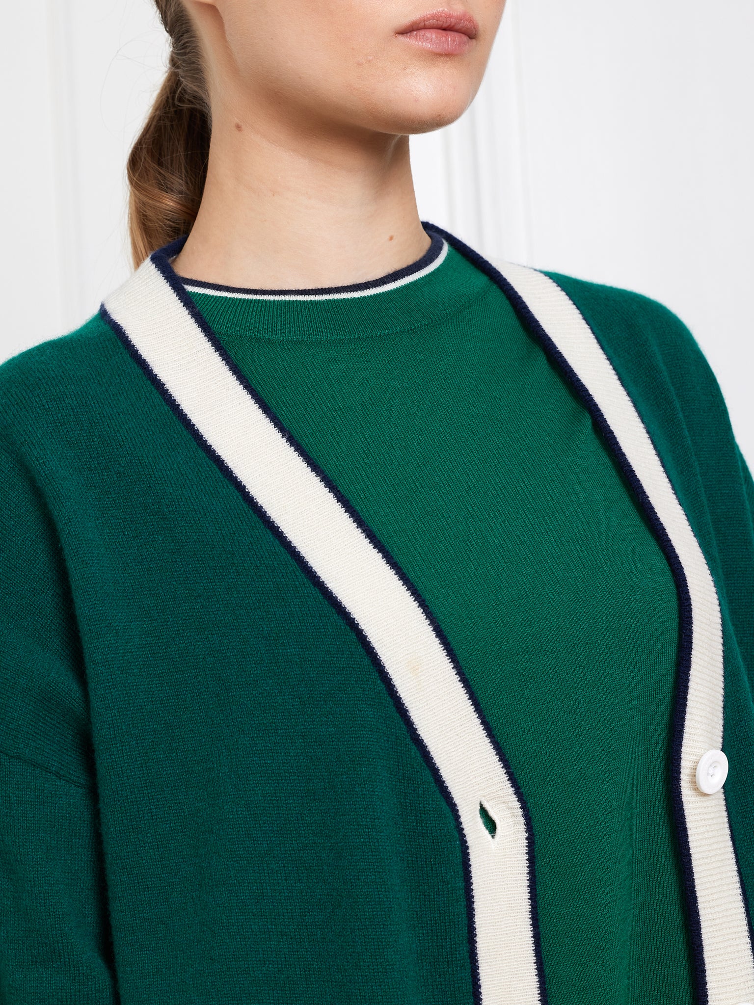 A person is elegantly styled in We Norwegians' Club Cardigan Women, an oversized cashmere piece with white and navy trim, layered over a green sweater. The exquisite knitwear is showcased from shoulders to mid-torso against a plain white wall.