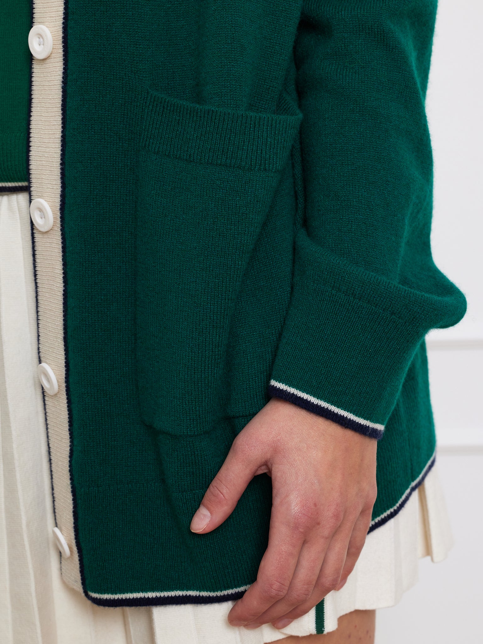 A person models the luxurious green "Club Cardigan Women" by We Norwegians, featuring white buttons and navy trim, with a hand on the pocket. They wear an oversized cream pleated skirt against a textured white backdrop.